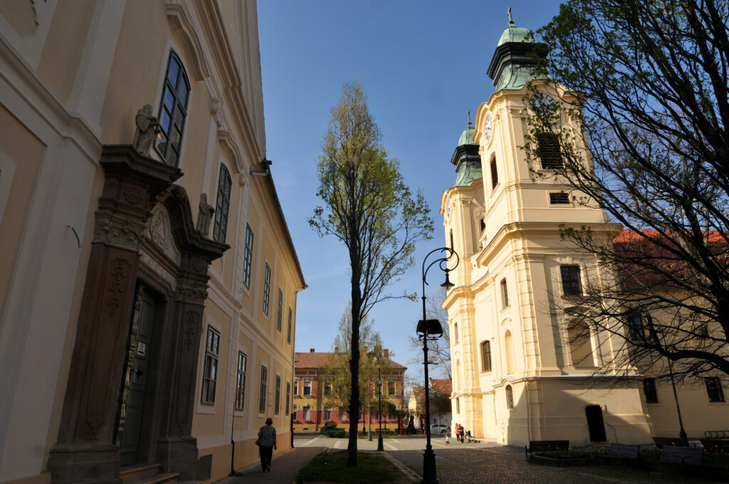 Utcaképi fotó a celldömölki kegytemplomról. A háttérben a templom világossárga épülete látszik két zöld sisakos, órás tornyával. Az előtérben balra egy másik épület barokk stílusú, szoborral díszített sarka és oszlopa látható. A két épület között kövezett tér, zöld fák, egy kovácsoltvas jellegű utcai lámpaoszlop és egy pad helyezkedik el.