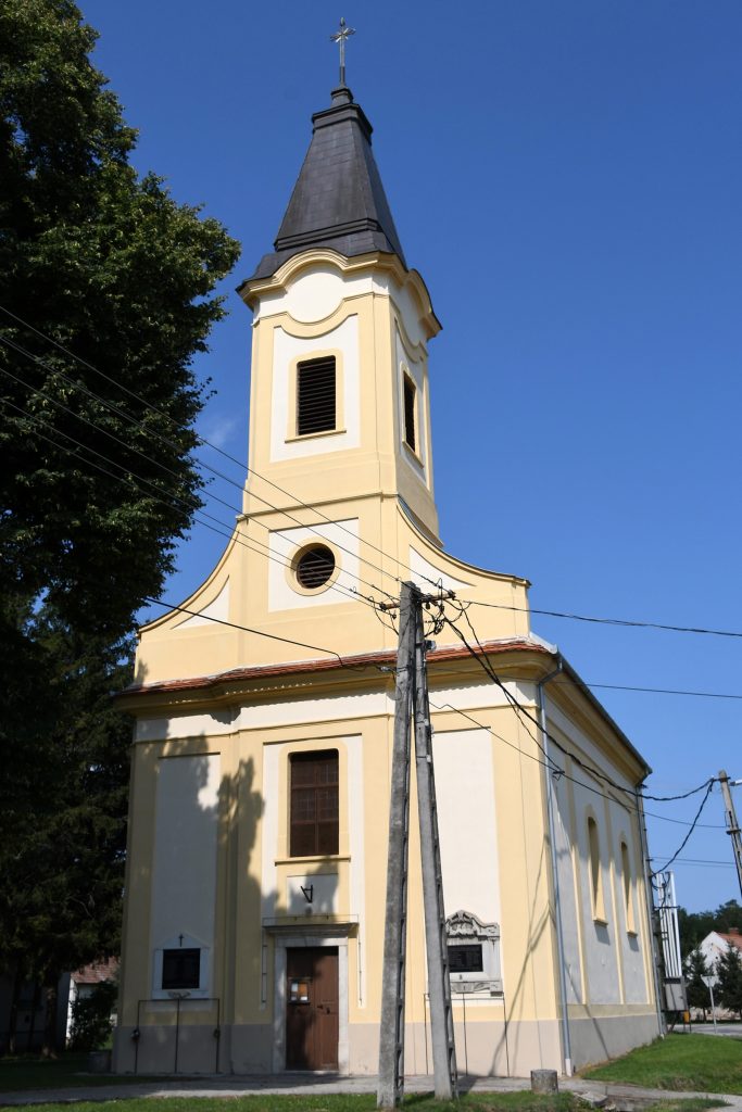 Színes fotó az izsákfai római katolikus templomról. A fehér falú, klasszicizáló stílusú templom sárga keretezésű ablakokkal és egy zömök, gúlasisakos toronnyal rendelkezik. Az épületet ápolt zöld gyep és alacsony kerítés veszi körül.