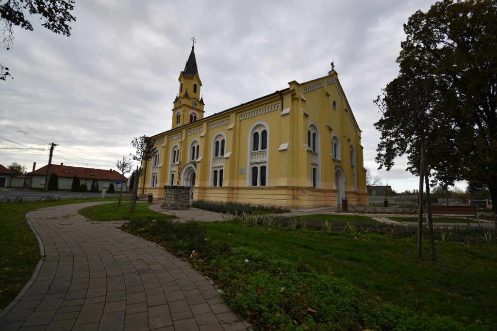 A nemesdömölki evangélikus templom sárga épülete egy gondozott parkkal, padokkal és virágágyásokkal az előtérben, felhős égbolt alatt.