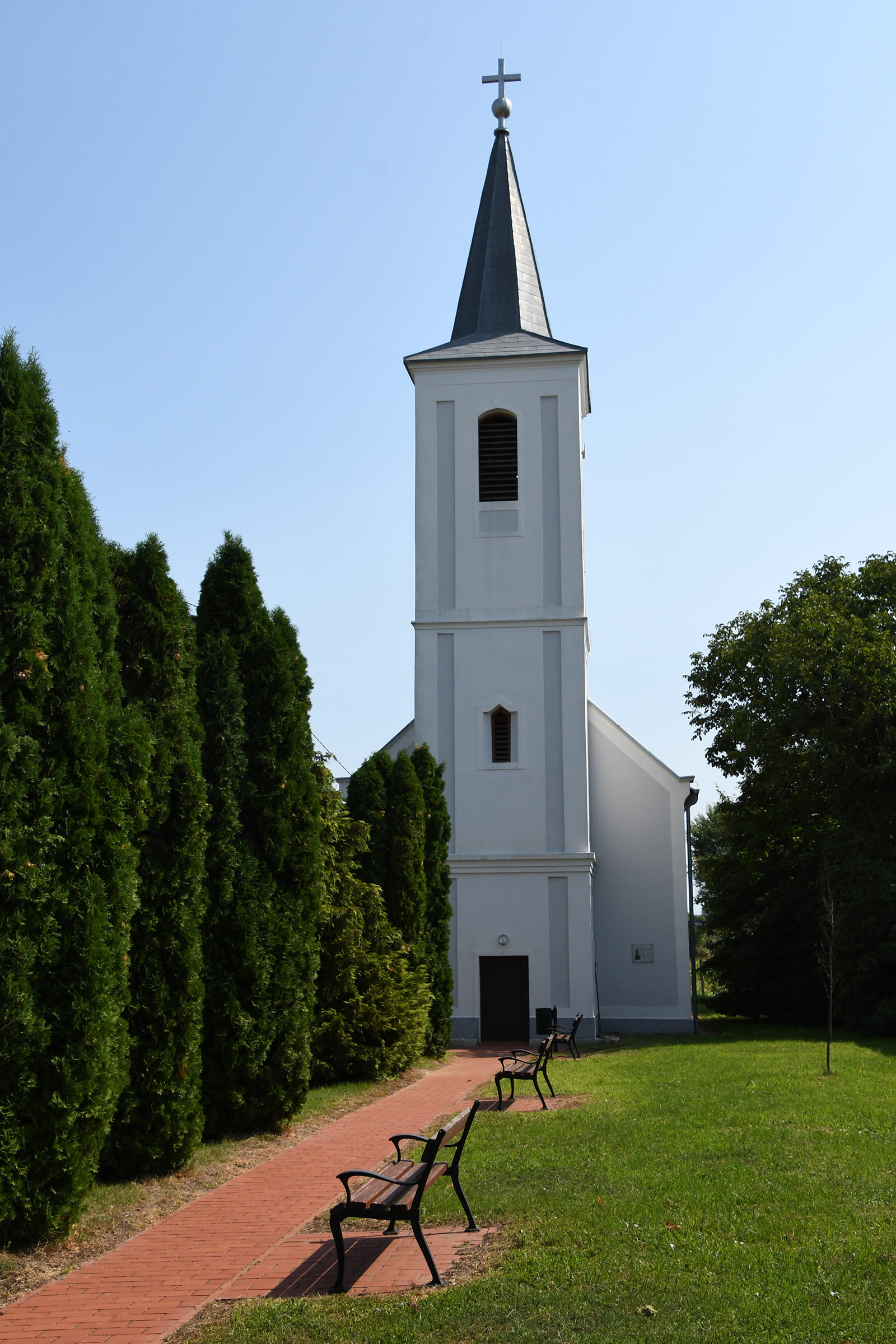 „A nemeskocsi evangélikus templom épülete látható egy vörös téglás sétány végén. A templom hófehér homlokzatú, karcsú tornya sötétszürke, gúla alakú sisakban végződik, melynek csúcsán kereszt látható. A sétány bal oldalán magas, zöld tujasor húzódik, a jobb oldalon pedig gondozott pázsit terül el padokkal. Az égbolt tiszta kék, a napfény élesen világítja meg a templomtorony oldalát.”
