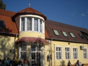Szent Benedek Katolikus Általános Iskola épülete Celldömölkön. A sárga színű épület hátsó bejárataegy kétszintes, kör alakú toronyrész, melynek tetején egy kereszt látható. A bejáratot egy rácsos fém szerkezet keretezi.