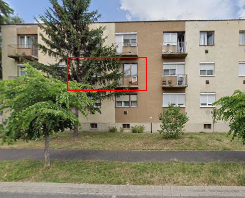 Utcai nézet egy többszintes, sárga és bézs homlokzatú társasházról. Az előtérben zöld fák és füves terület látható, a ház előtt járda húzódik. A kép közepén egy piros téglalap jelöli az első emeleti erkélyes lakást, amely részben egy fenyőfa ágai mögött található. Eladó lakás - Budai Nagy Antal u. 8/B. 1/3.