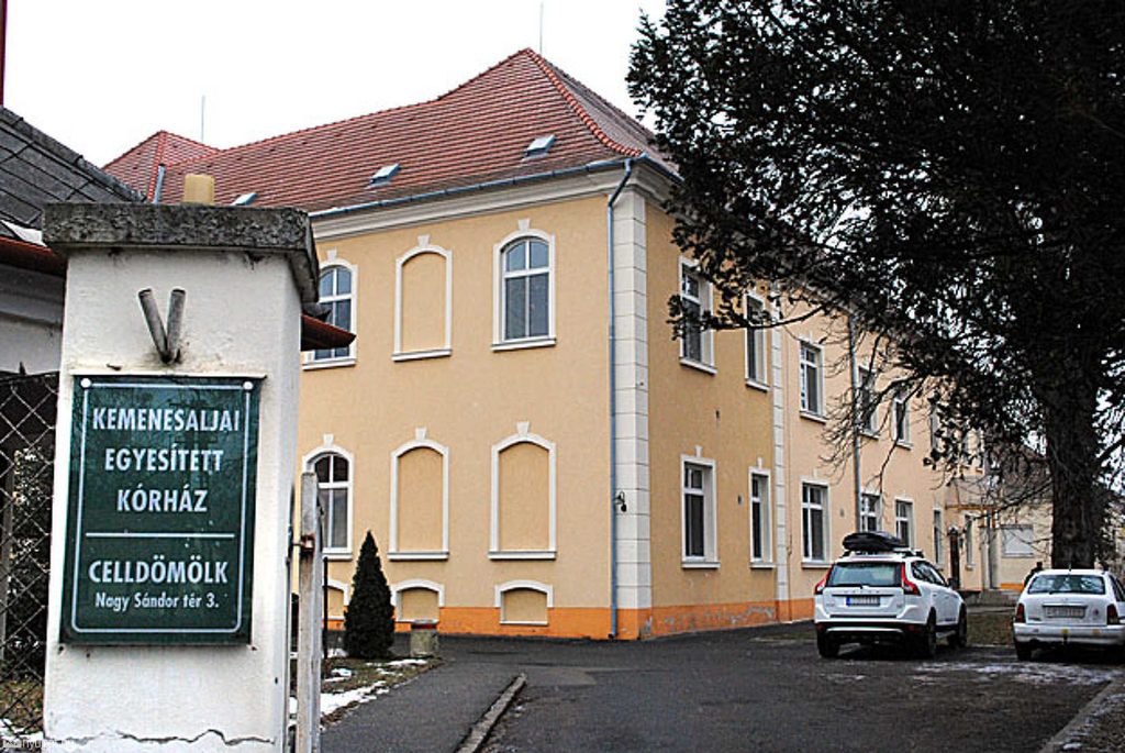Fotó a celldömölki Kemenesaljai Egyesített Kórház épületéről. Az előtérben egy fehér oszlopon zöld tábla látható a kórház nevével és címével (Nagy Sándor tér 3.). A háttérben a kórház kétszintes, sárga falú, vörös tetős épülete és parkoló autók látszanak.